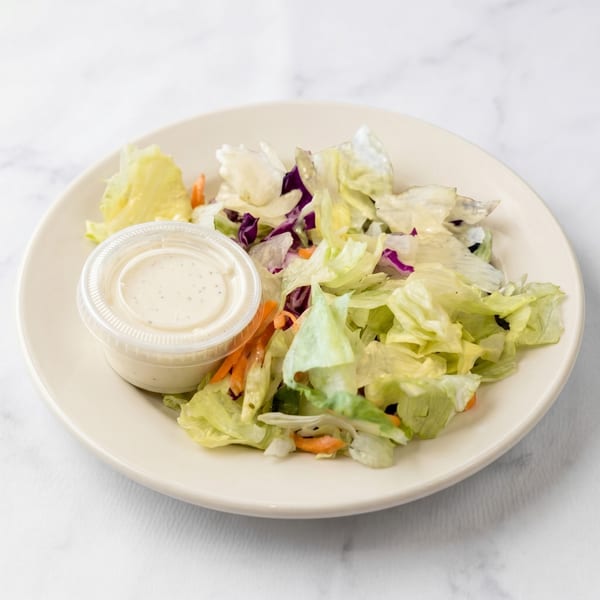 Side salad with mixed greens, shredded carrots, and a cup of ranch dressing on a white plate.